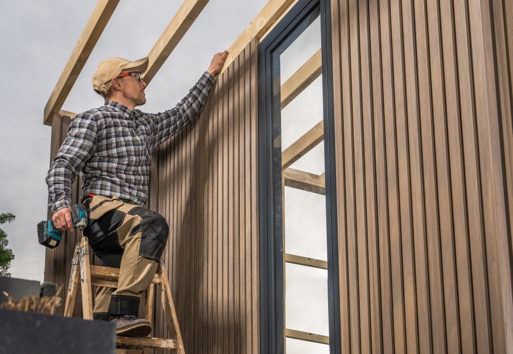 Experienced Caucasian Woodworker with Cordless Drill Driver in His Hands Building Modern Backyard Garden Shed. Composite Material Walls.