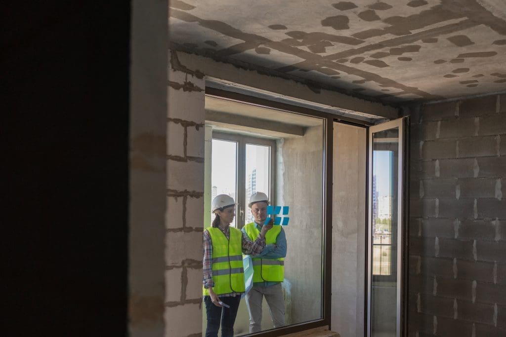 two engineers in protective vests and white helmets glue stickers on the window and discuss the construction plan of the facility or the scheme. European male and female engineers perform fire safety design work